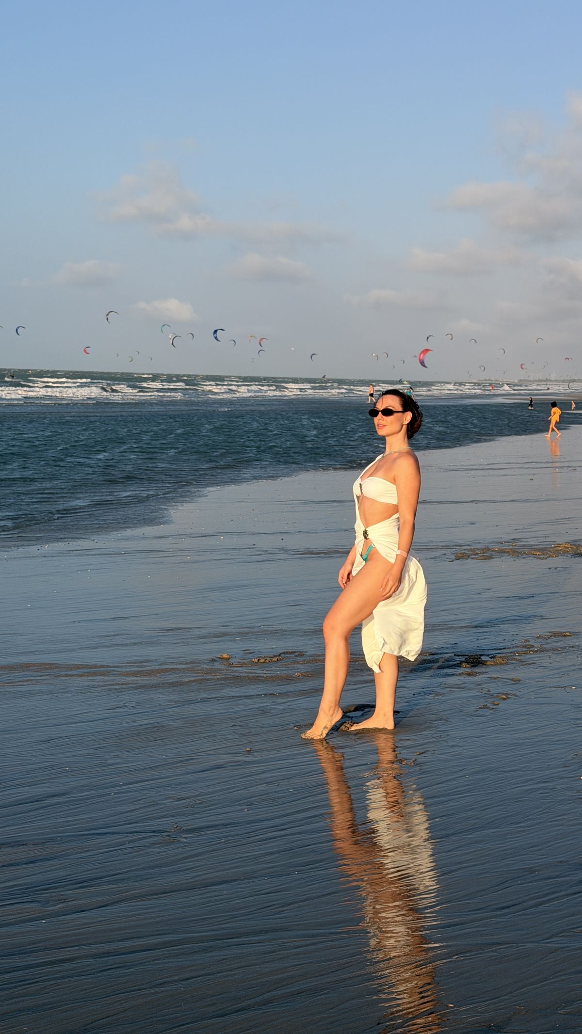 Lidiane Grigoletto na beira-mar da Praia do Preá na maré baixa, com kites no céu