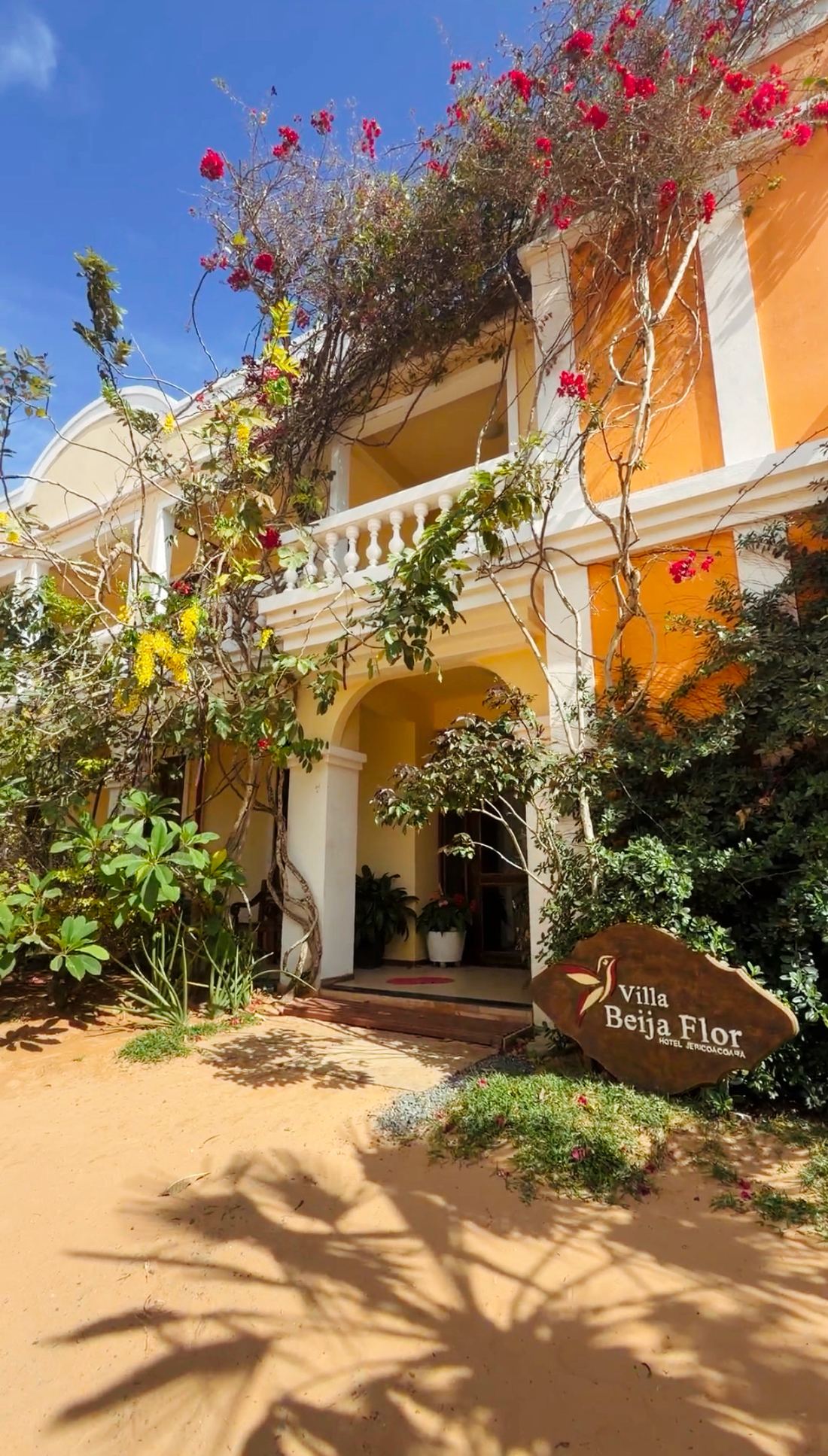 Entrada do Hotel Villa Beija Flor em Jericoacoara, com fachada amarela, buganvílias e a placa na areia